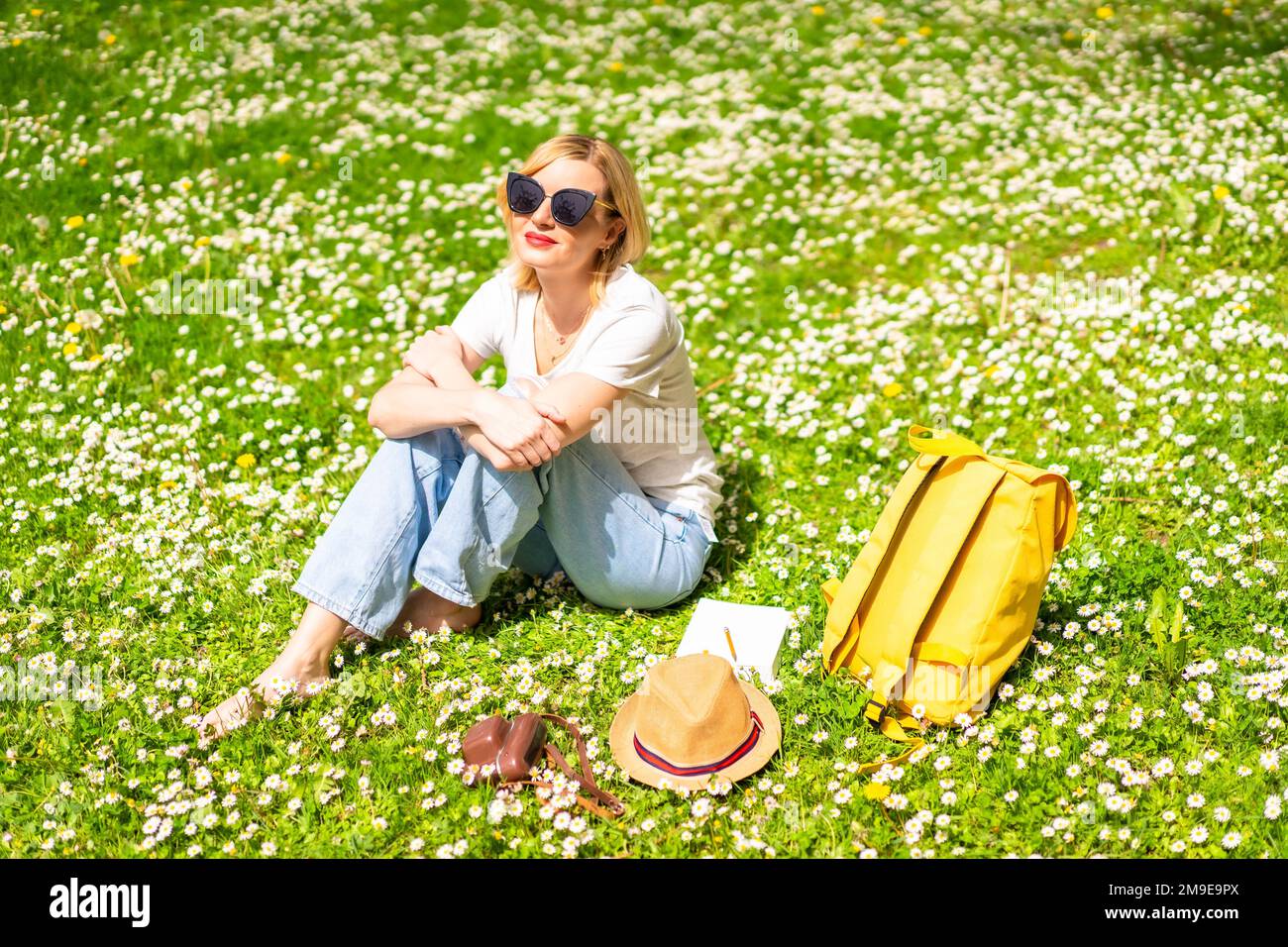 Una giovane bionda in un cappello che si distenderà e respirerà puro lehaire in primavera in un parco della città, la natura, seduto sull'erba accanto alle margherite Foto Stock