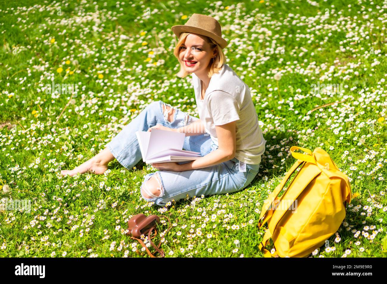 Una giovane bionda in un cappello scrivere poesie in un taccuino in primavera in un parco della città, la natura, seduto sull'erba accanto alle margherite Foto Stock