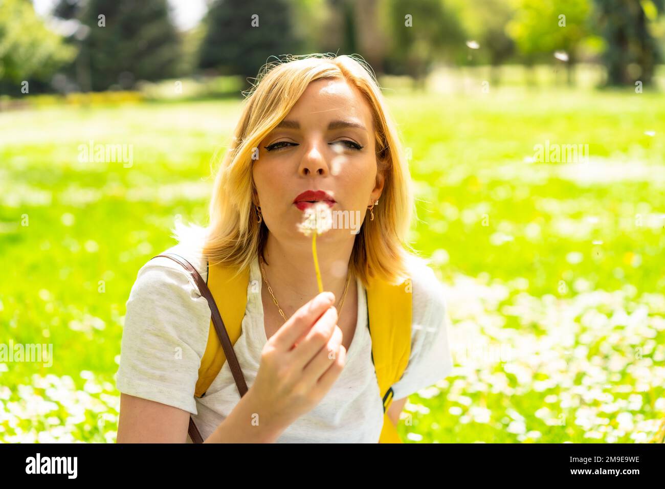 Una donna bionda sorridente che soffia sulla pianta del dente di leone, indossando un cappello e occhiali da sole seduti sull'erba in primavera accanto alle margherite in un parco nel Foto Stock