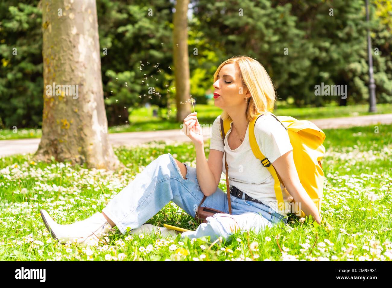 Una donna bionda che soffia sulla pianta del dente di leone in un parco della città, indossando un cappello e occhiali da sole seduti sull'erba in primavera accanto alle margherite Foto Stock