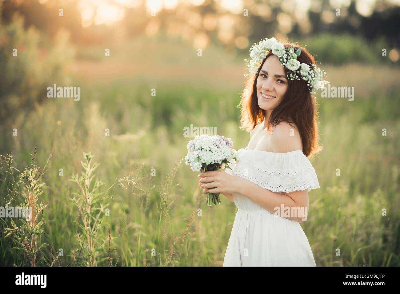 Ragazza felice caucasica in un vestito bianco con un bouquet di fiori e una corona d'estate al tramonto sul campo. Unità e armonia con la natura Foto Stock