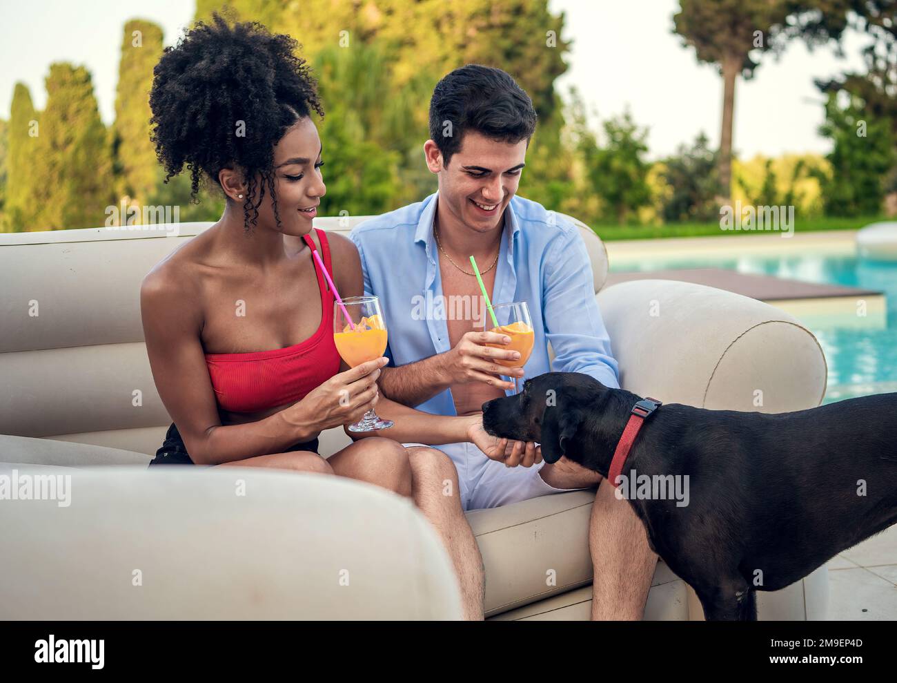 Una giovane coppia interracial si siede su un divano da giardino in una lussuosa villa o resort. Tenere bicchieri di succo d'arancia e stanno sorridendo mentre coccolare un cane - D Foto Stock