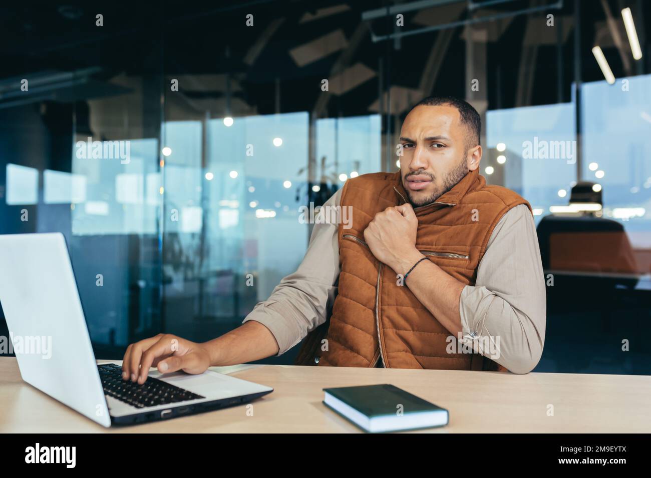 Uomo afroamericano che congela in ufficio, non riscalda sul posto di lavoro, uomo in outerwear seduto alla scrivania con un computer portatile al lavoro. Foto Stock