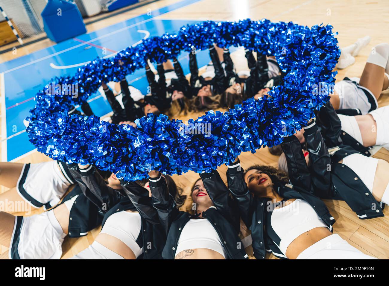 Cheerleaders che si adagia su un campo da basket tenendo i loro pom-pom blu e lucenti in cerchio. Foto di alta qualità Foto Stock