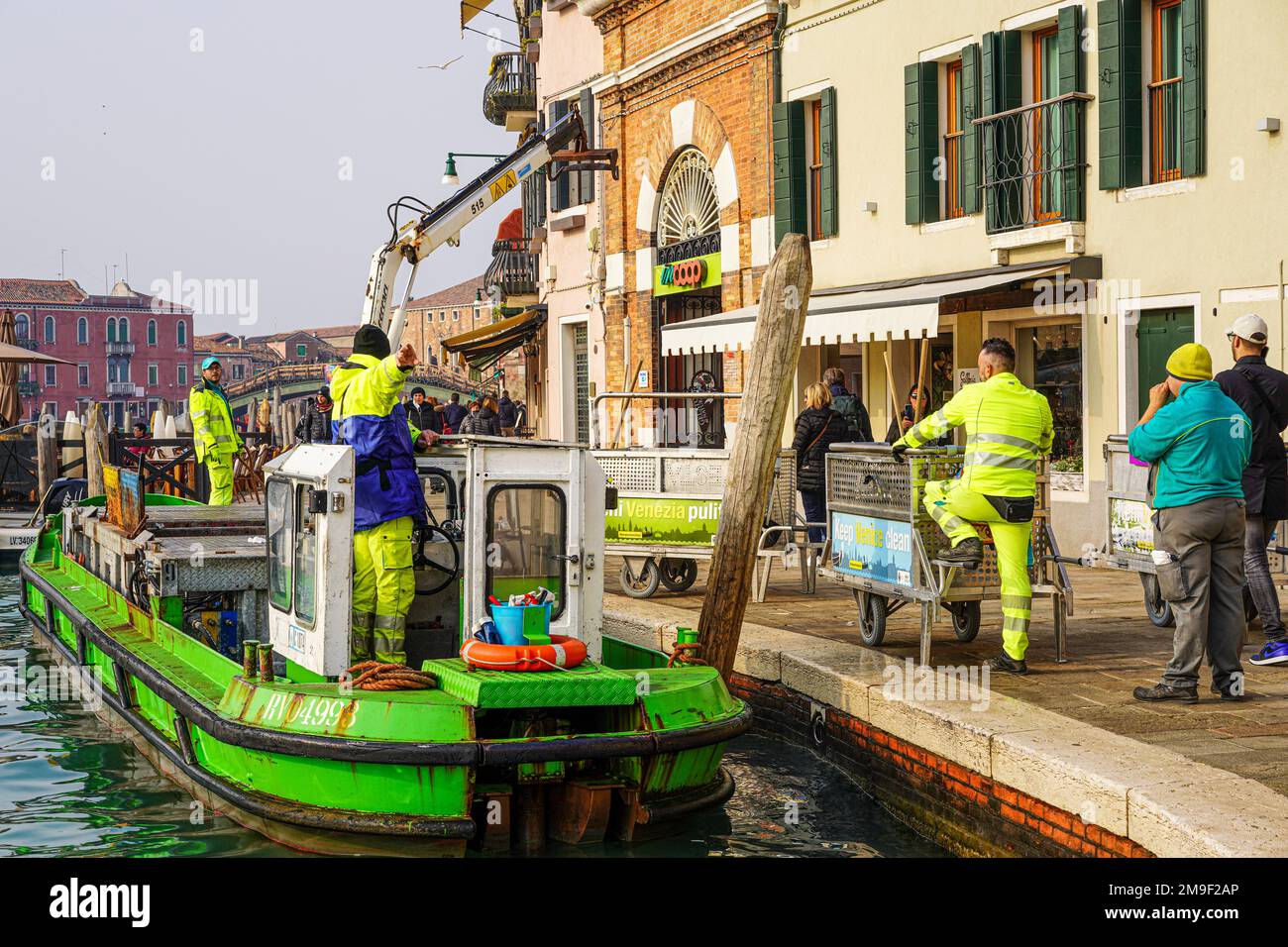 Una nave di rifiuti si ferma in un canale. Gli uomini in giacche gialle spingono i contenitori con i rifiuti alla nave, aspettano finché la gru della nave li porta sulla nave. Foto Stock