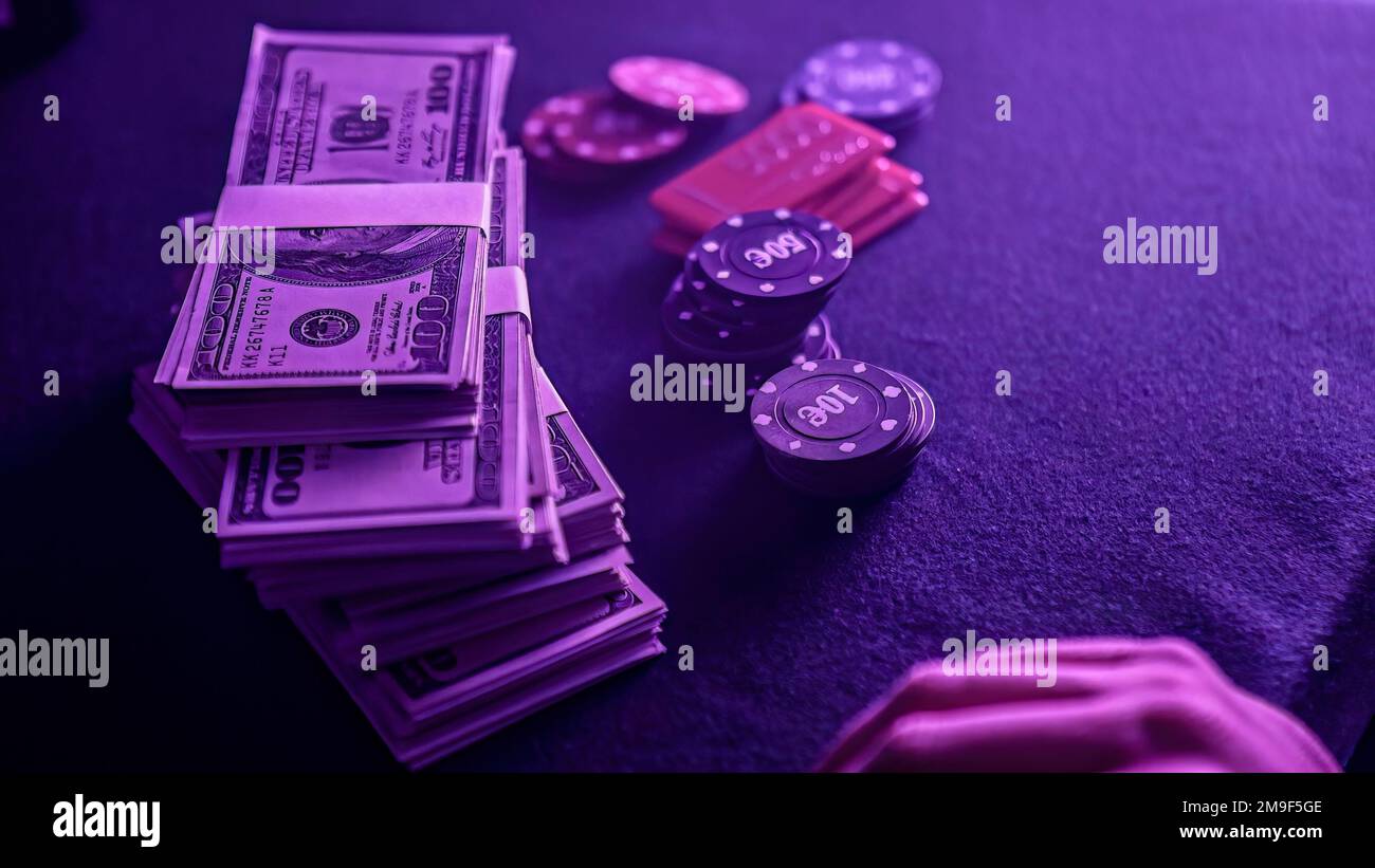 Pila di chip di poker in una gamma di colori e una bacchetta di dollari sul tavolo da gioco in un casinò scarsamente illuminato. Le fiche e i soldi suggeriscono un gioco di poker Foto Stock