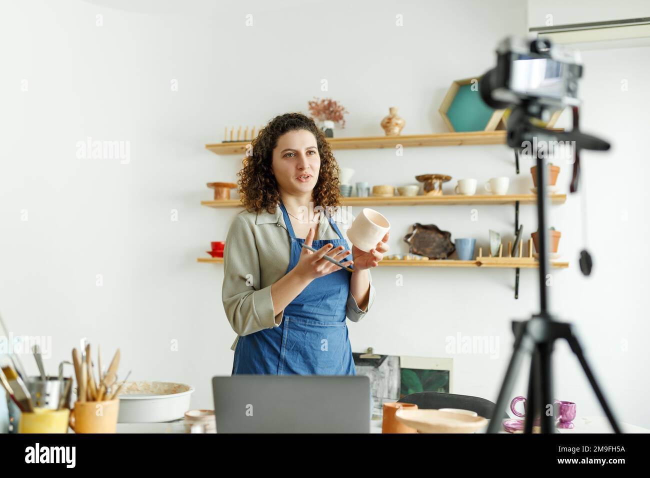 Femmine vasaio fare un vlog, registrazione corso on-line, lezioni nel suo studio con scaffale di terracotta sullo sfondo. Foto Stock