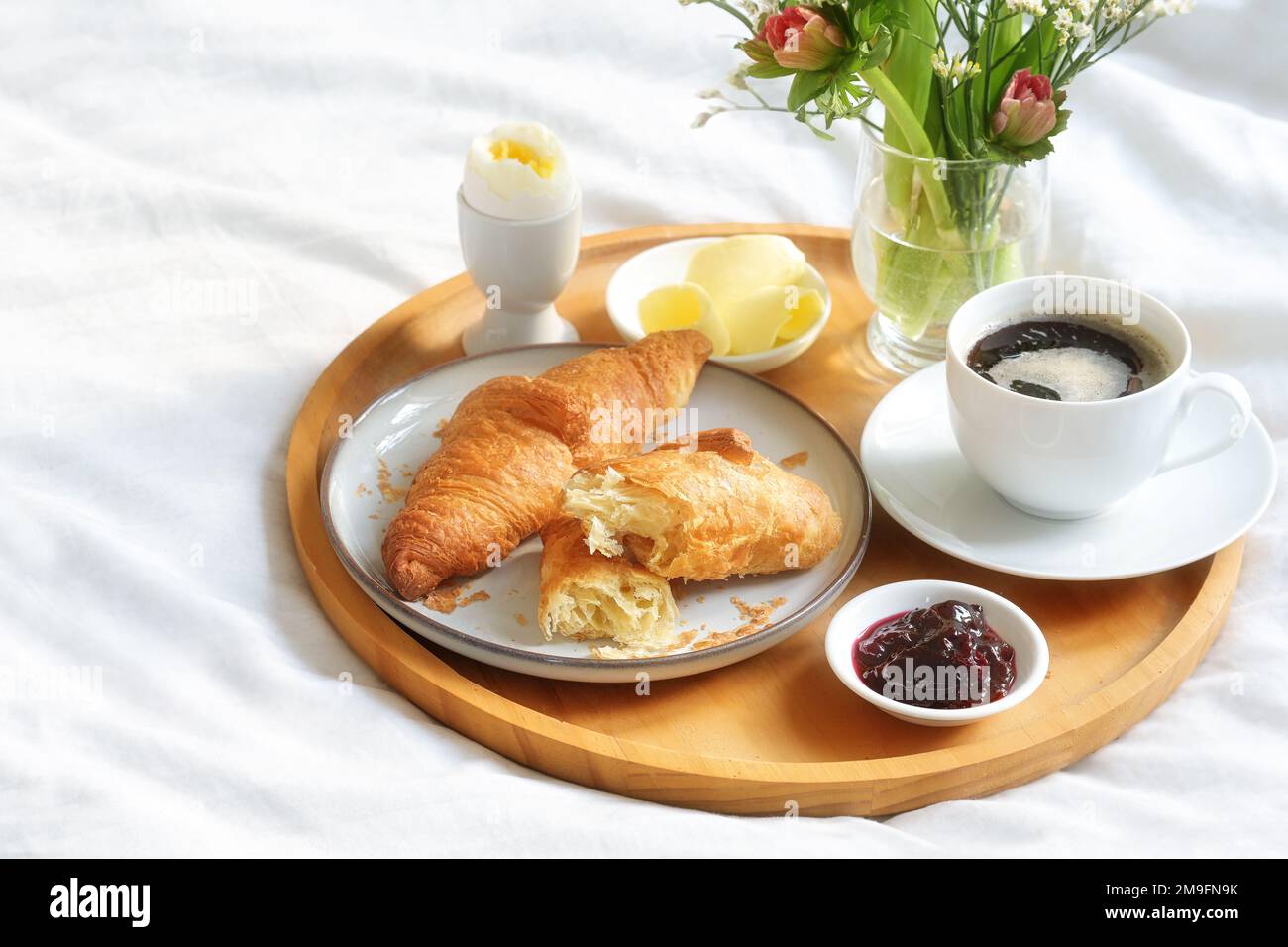 Piccola colazione con croissant, caffè, marmellata e uova su un vassoio di legno con un bouquet di fiori su un letto bianco coperto di Domenica, compleanno, di San Valentino o. Foto Stock