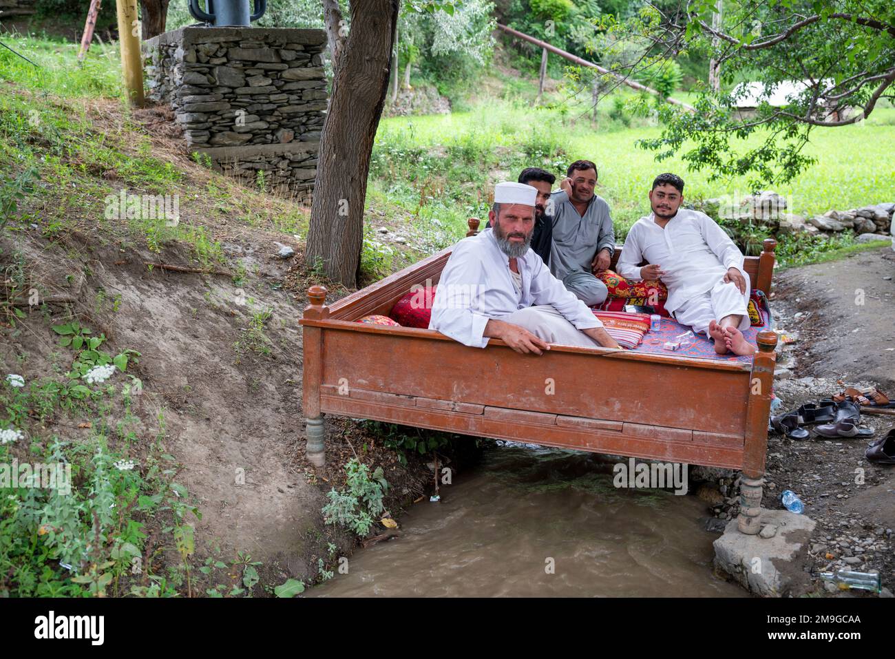 Un gruppo di uomini Kalash adulti che si rilassano su una panchina di legno sopra un ruscello, Bumburet Valley, Pakistan Foto Stock