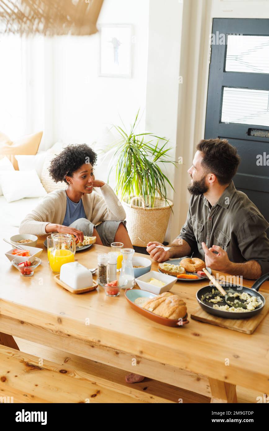 Vista dall'alto della giovane coppia biraciale che parla e fa colazione mentre si siede al tavolo da pranzo Foto Stock