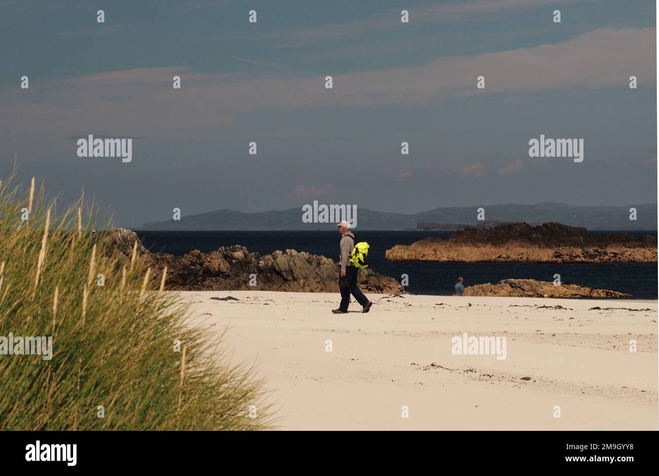 Un uomo 60+ che cammina sulla White Strand della spiaggia di Monk, Iona, Scozia UK, mostrando la sabbia bianca e il mare blu profondo e le isole sullo sfondo Foto Stock