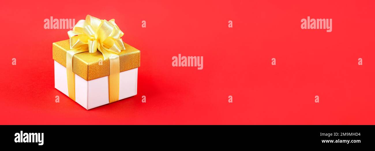 Confezione regalo dorata con fiocco a nastro satinato su sfondo rosso per una vacanza. Buon San Valentino, festa della mamma e auguri di compleanno. Banner Foto Stock