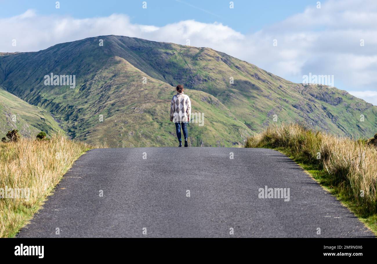 Una donna aggraziata che cammina lungo la strada per le montagne di Connemara, County Galway, Irlanda Foto Stock