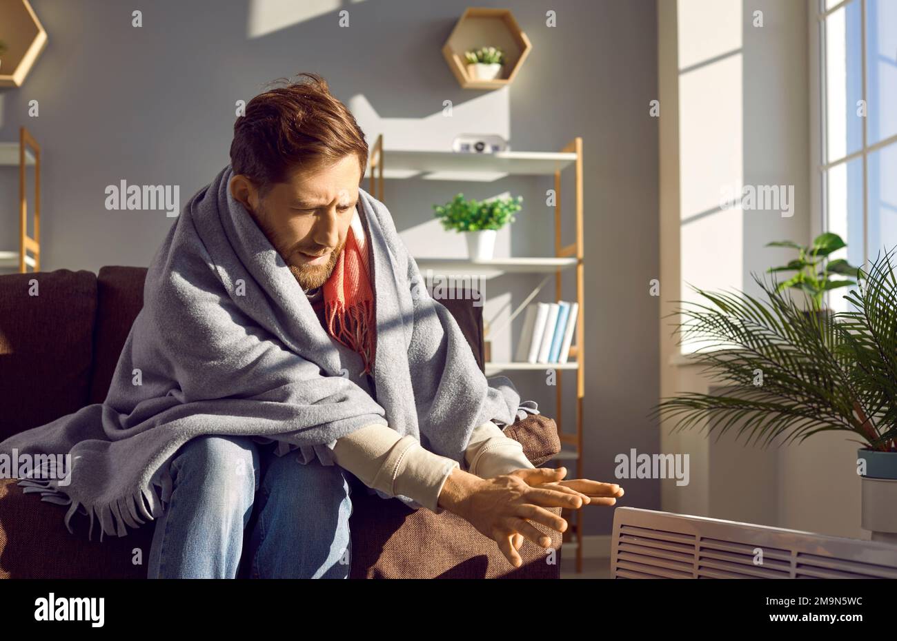 Giovane uomo congelato seduto avvolto in coperta a casa e cercando di riscaldare le mani sul riscaldatore elettrico Foto Stock