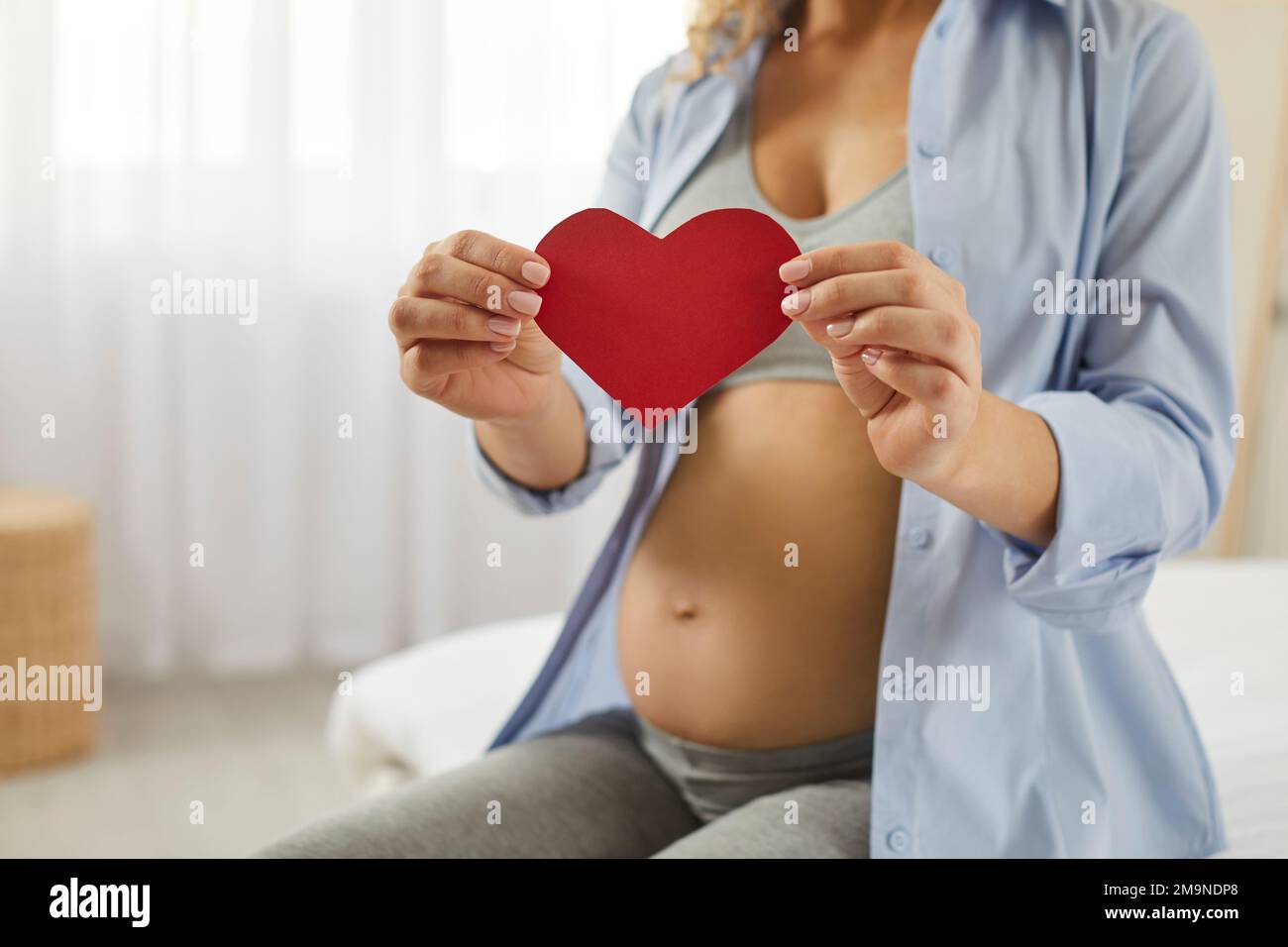 Giovane madre amorevole felice che si aspetta un bambino che tiene un cuore di carta nelle sue mani Foto Stock
