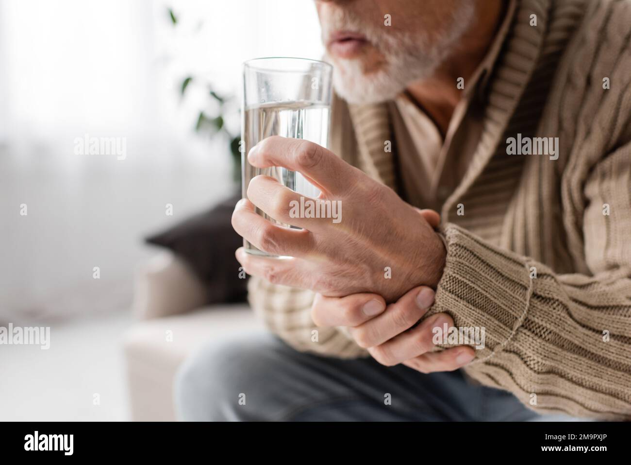vista ritagliata di un uomo anziano con parkinsonismo tenendo un bicchiere d'acqua in mani tremanti mentre si siede a casa, immagine stock Foto Stock