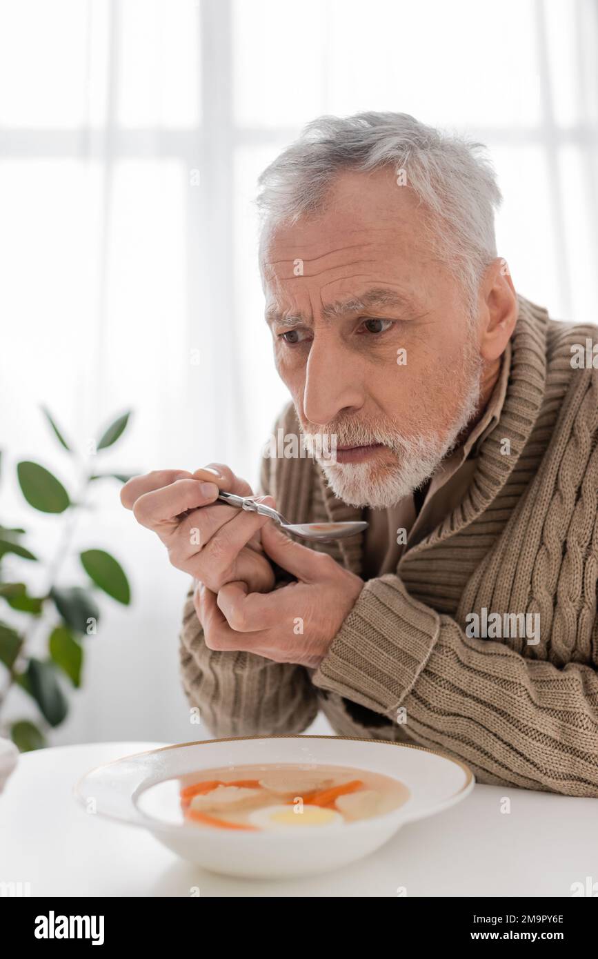 uomo depresso con sindrome parkinsoniana seduta con cucchiaio in mani tremanti vicino zuppa in cucina, immagine stock Foto Stock