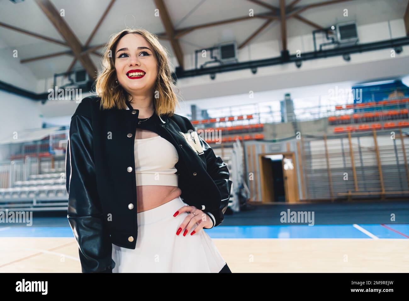 Scatto medio di giovane ragazza cheerleader in uniforme bianca e nera sorridente e in posa in sala sportiva. Foto di alta qualità Foto Stock