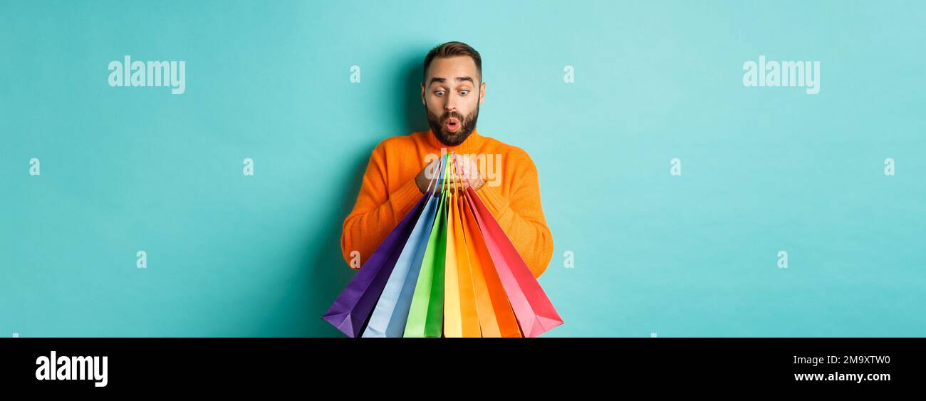 Uomo eccitato che guarda le borse della spesa con gli acquisti, in piedi divertito contro sfondo turchese Foto Stock