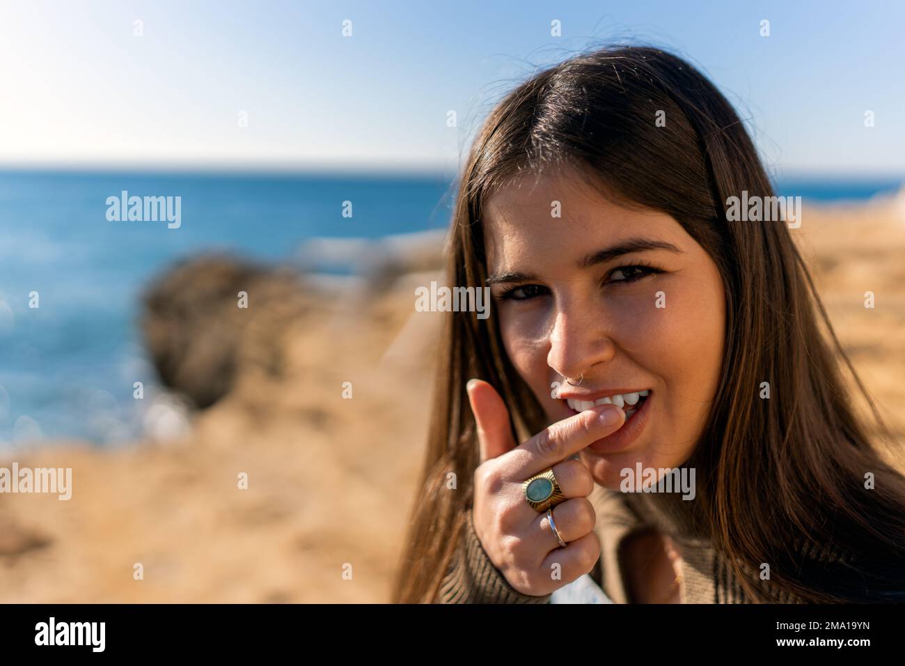 Donna etnica soddisfatta con i capelli lunghi che guardano la macchina fotografica mentre tocca i denti sulla spiaggia sabbiosa nella giornata di sole Foto Stock