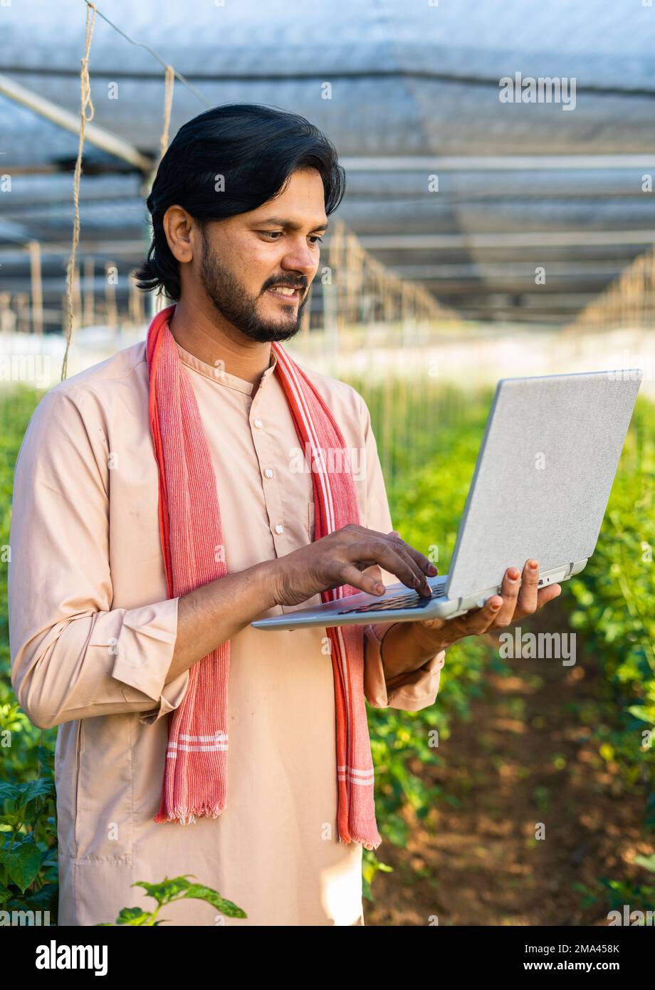 Immagine verticale di un giovane agricoltore felice impegnato a lavorare sul computer portatile in serra - concetto di agricoltura moderna, tecnologia e sviluppo o crescita. Foto Stock