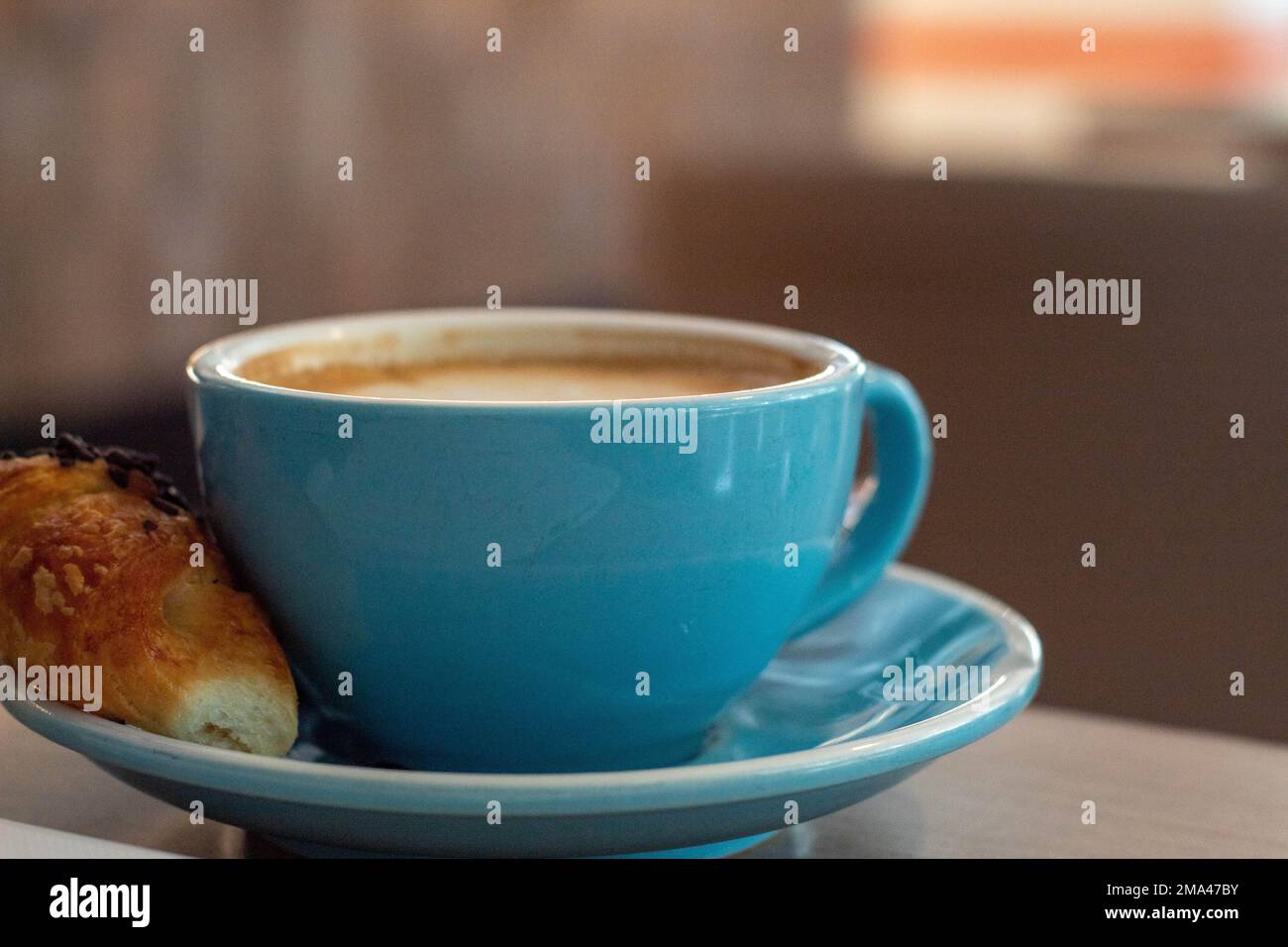 Caffè con latte in una grande tazza blu con un mini croissant mentre si telelavoro in una caffetteria ad Alicante con lo sfondo fuori fuoco. Foto Stock