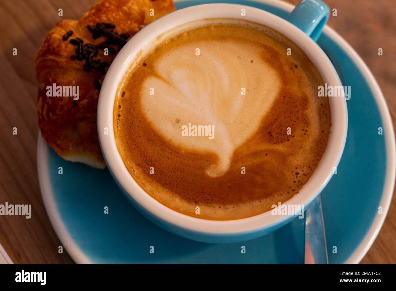 Caffè con latte in una grande tazza blu con un mini croissant mentre si telelavoro in una caffetteria ad Alicante con lo sfondo fuori fuoco. Foto Stock