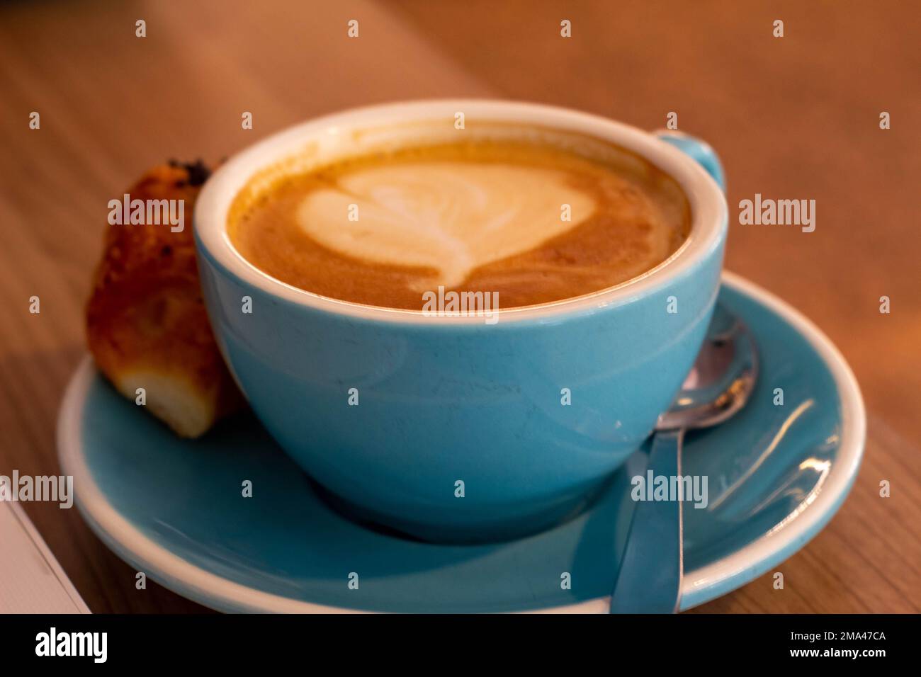 Caffè con latte in una grande tazza blu con un mini croissant mentre si telelavoro in una caffetteria ad Alicante con lo sfondo fuori fuoco. Foto Stock