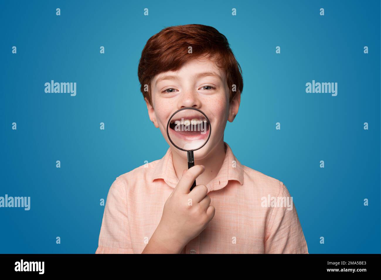 Foto di un ragazzino di zenzero candido positivo che tiene una lente d'ingrandimento che mostra i denti della fotocamera isolato su sfondo di colore blu Foto Stock