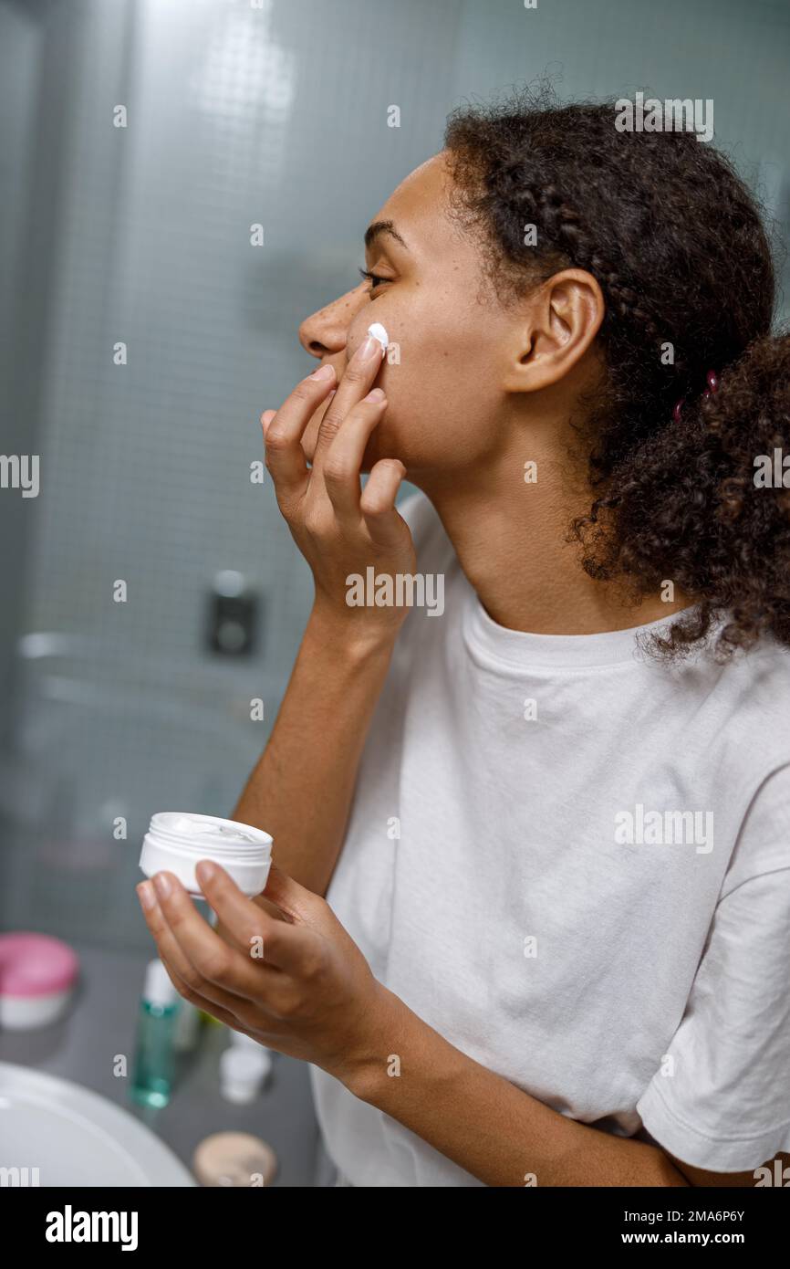 Donna Africana che applica crema idratante in piedi dietro lo specchio nella stanza da bagno domestica. Procedura di bellezza Foto Stock