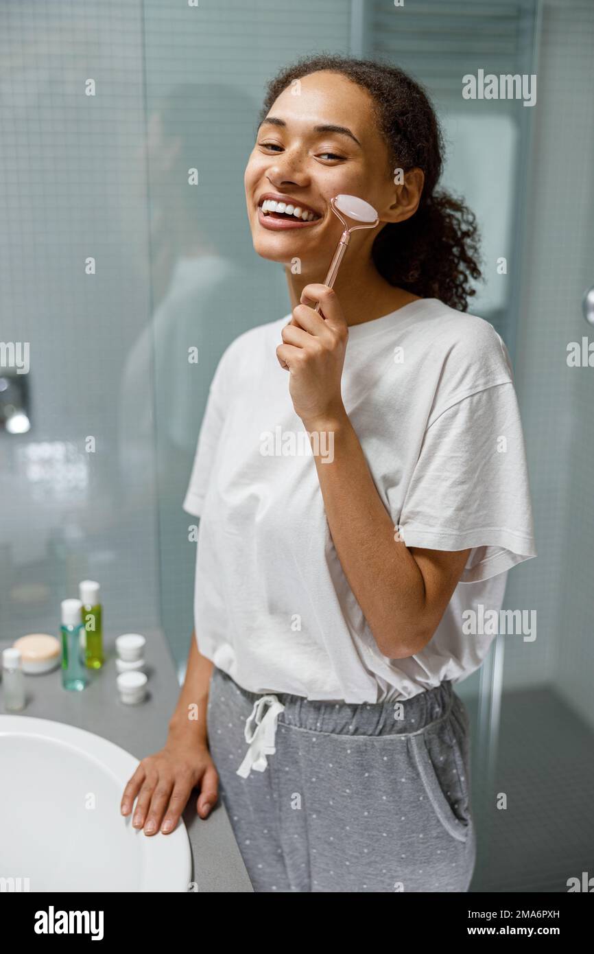 Donna sorridente che fa il massaggio del viso usando il rullo con il quarzo in bagno. Concetto naturale di cosmetica Foto Stock