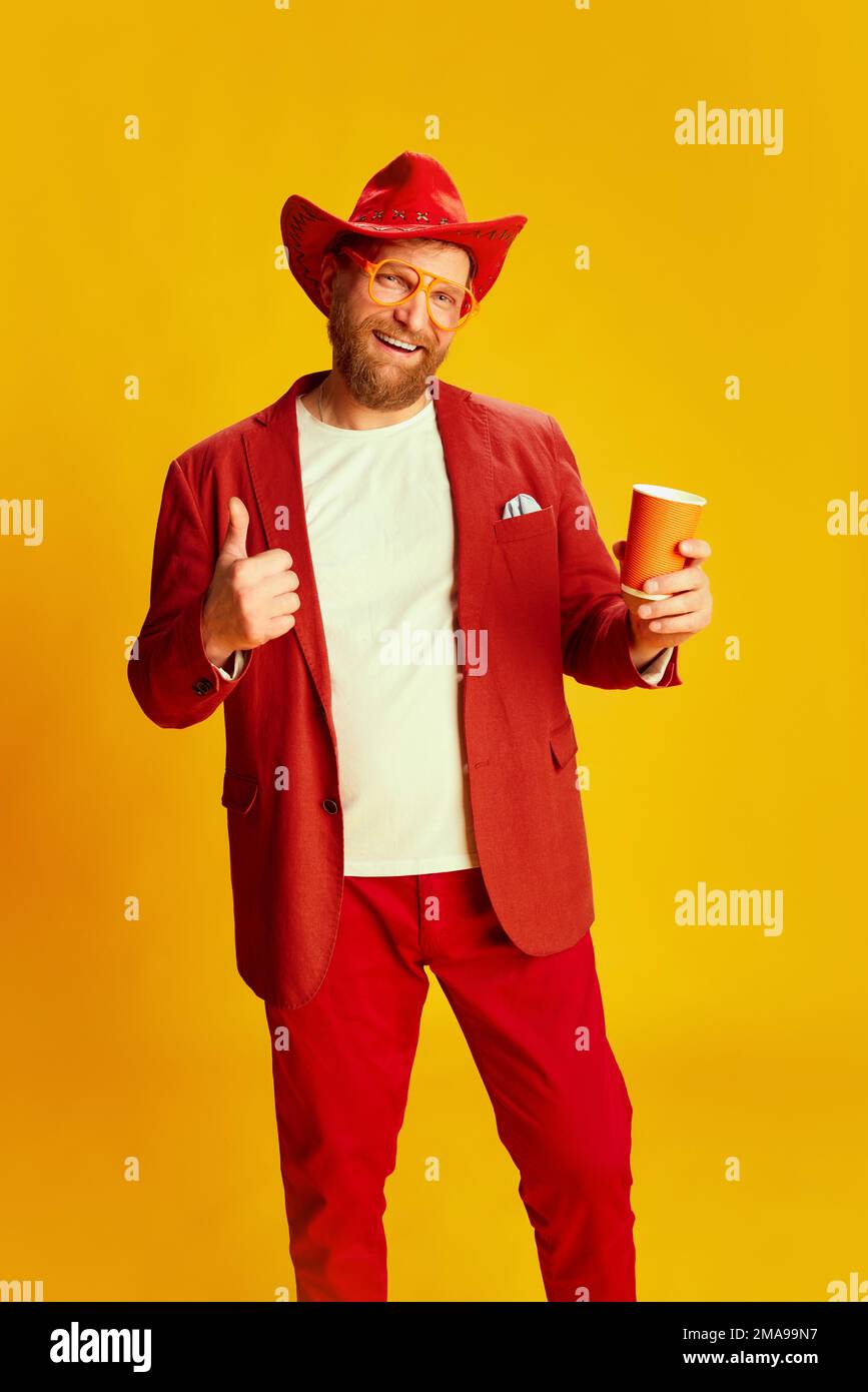Uomo alla moda, hipster con costume rosso brillante e cappello da cowboy che si posa su sfondo giallo. Concetto di stile , moda, umore felice, positivo Foto Stock