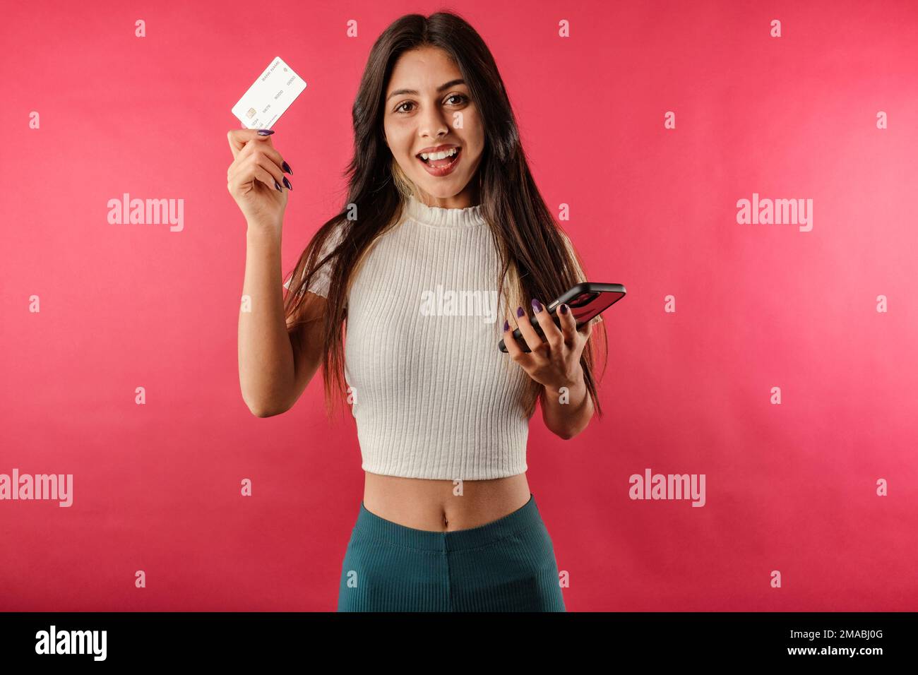 Felice donna brunette isolato su sfondo rosso con una carta di credito e un telefono sembra overjoyed e guarda la fotocamera. Vedere un campo incredibile Foto Stock