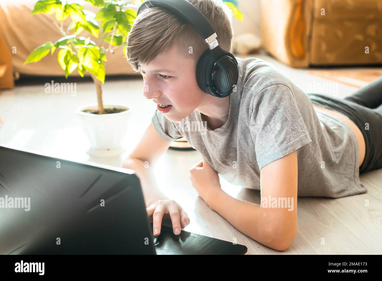 ragazzo con cuffie wireless per gamer si trova sul pavimento e gioca a giochi online. il bambino impara i programmi a distanza a casa nel giardino d'inverno vicino al fiore in vaso Foto Stock