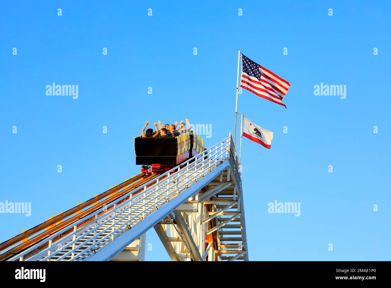 I turisti cavalcano le montagne russe che si avvicinano alla cima con la bandiera dello stato della California e stelle e strisce. Santa Monica, California, Stati Uniti Foto Stock