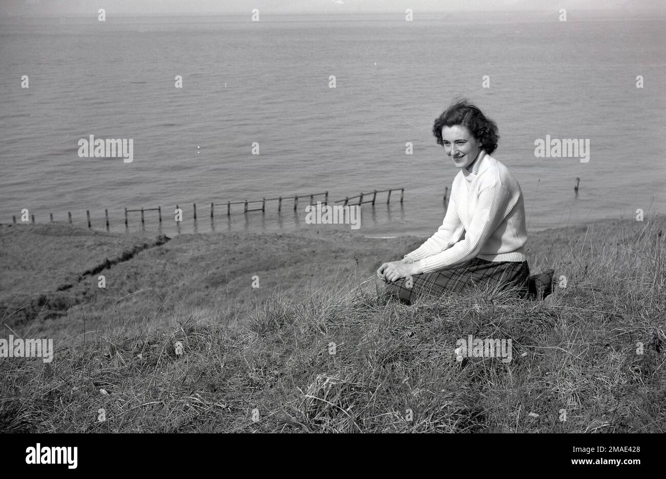1950s, storica, una giovane donna attraente, metà anni ventenni, indossando un top in cotone roll-neck e gonna a motivi tartan seduta per una foto su una collina erbosa che domina il mare, Inghilterra, Regno Unito. Foto Stock
