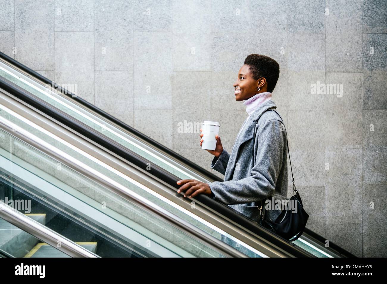 Vista laterale di un'elegante donna afro-americana in un elegante cappotto e maglione. Lei è con una tazza di caffè e una borsa sorridente e cavalcando le scale in movimento in città Foto Stock