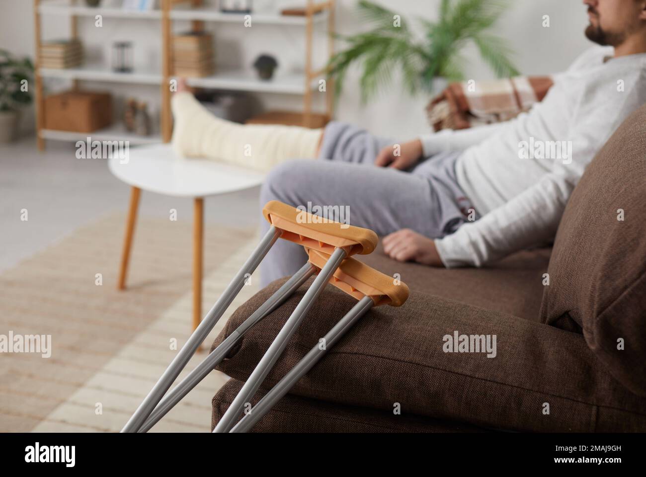 Primo piano di stampelle appoggiate sul divano, e un uomo con una gamba rotta seduto sullo sfondo Foto Stock