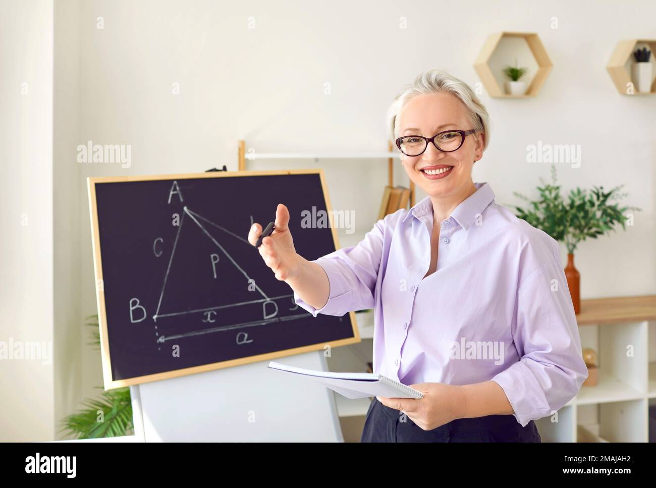 Allegra insegnante di donna dai capelli grigi o tutor privato si trova in una lavagna con disegni geometrici Foto Stock