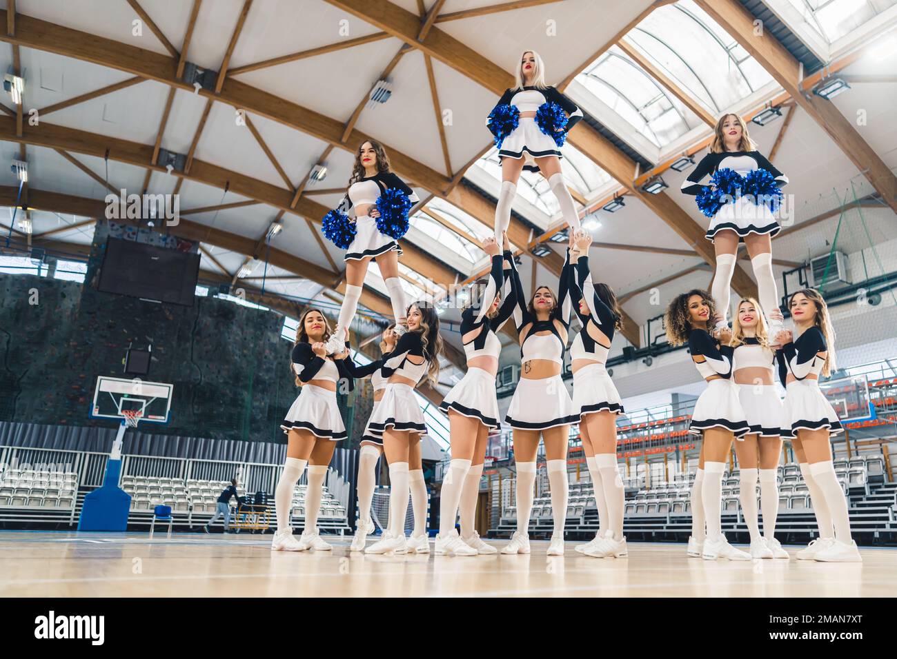 Vista dal basso della squadra cheerleading in divise a due colori facendo una figura ginnica mentre tiene tre membri in su. Tre membri della squadra che tengono in mano pom blu lucenti. Foto di alta qualità Foto Stock