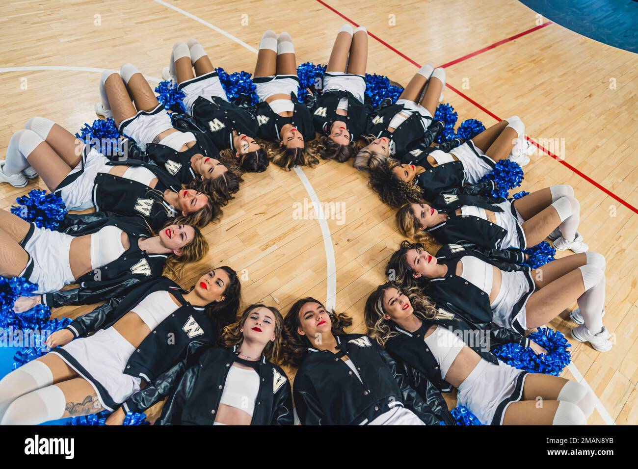 Cheerleading team in uniformi adagiato su un campo da basket in un cerchio con pom blu lucido nelle loro mani. Foto di alta qualità Foto Stock