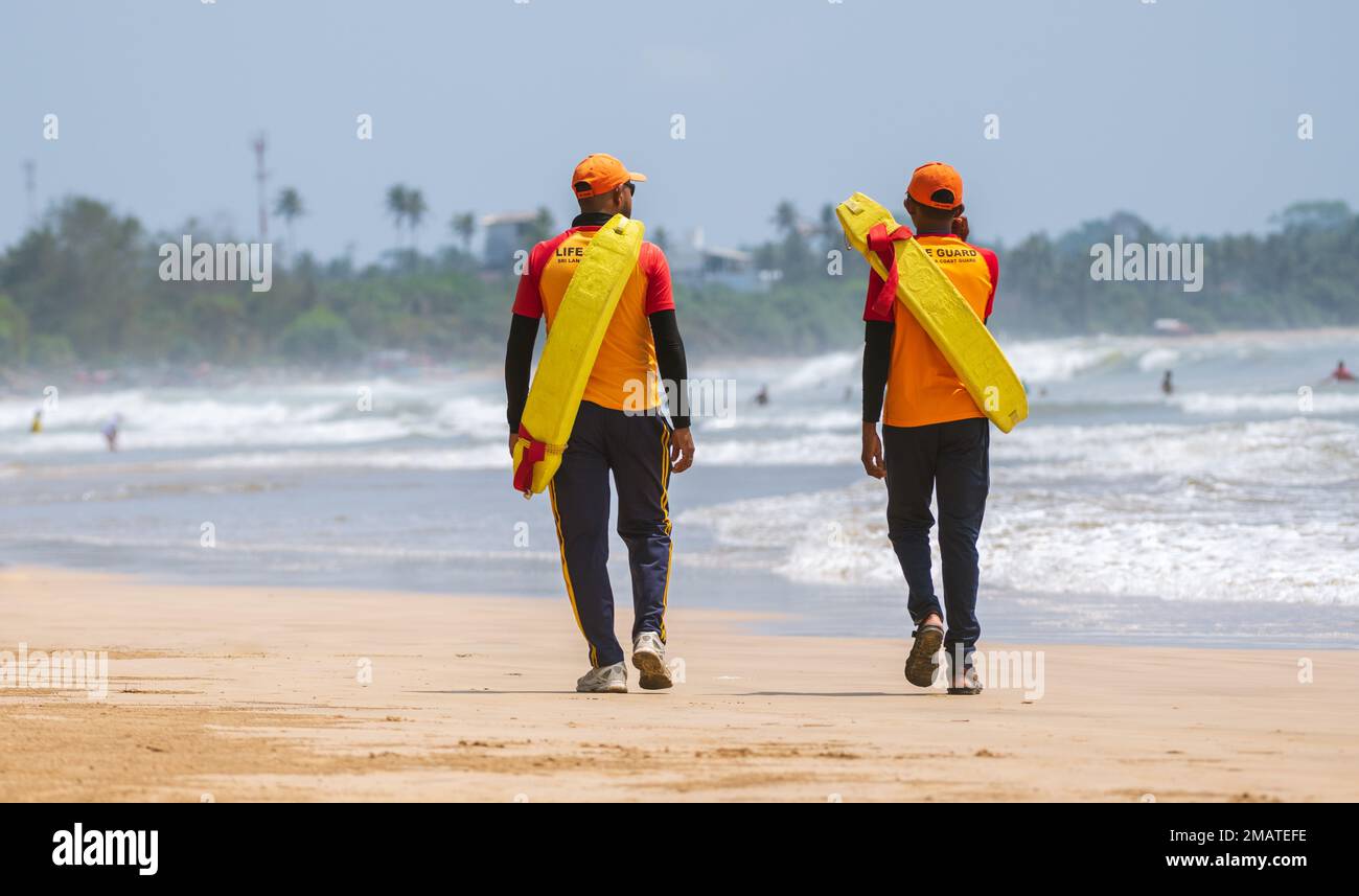 Due bagnini in servizio presso la spiaggia tropicale, entrambi trasportano tubi di salvataggio sulle spalle e camminano sulla spiaggia di sabbia tropicale a Weligama. Foto Stock