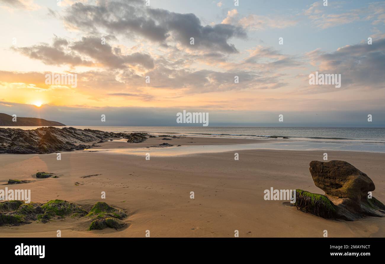 Alba, con rocce sulla spiaggia, e sabbia dorata, e la luce dorata riflessa nella sabbia bagnata, creando una tranquilla scena. Foto Stock