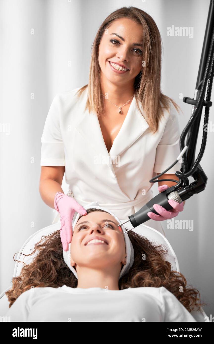 Ritratto di donna sorridente cosmetologa che esegue la resurfacing della pelle laser. Cliente felice in previsione del ringiovanimento della pelle dopo la procedura importante. Foto Stock