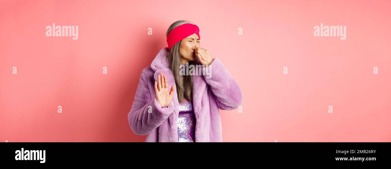 Donna matura asiatica disgustata in trendy cappotto e abito viola invernale, chiuso il naso e mostrare stop, gesto di rifiuto, odore qualcosa di disgustoso, in piedi Foto Stock