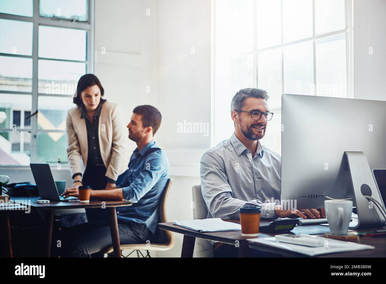 Ufficio, computer e uomo d'affari con i suoi colleghi che parlano o discutono in background. PC, successo e professionista maturo dipendente maschio Foto Stock