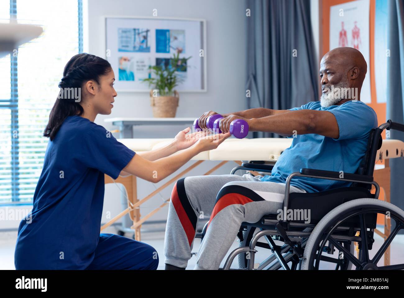 Fisioterapista femminile diversificata che aiuta il paziente maschio anziano nell'esercitazione della sedia a rotelle con i dumbbells Foto Stock