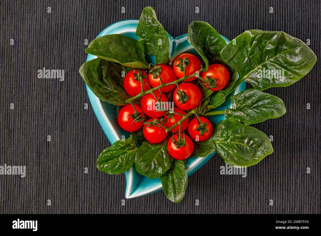 Pomodori ciliegini rossi su un ramo e foglie di spinaci verdi in una ciotola blu a forma di cuore un cuore su una tovaglia tessile nera Foto Stock