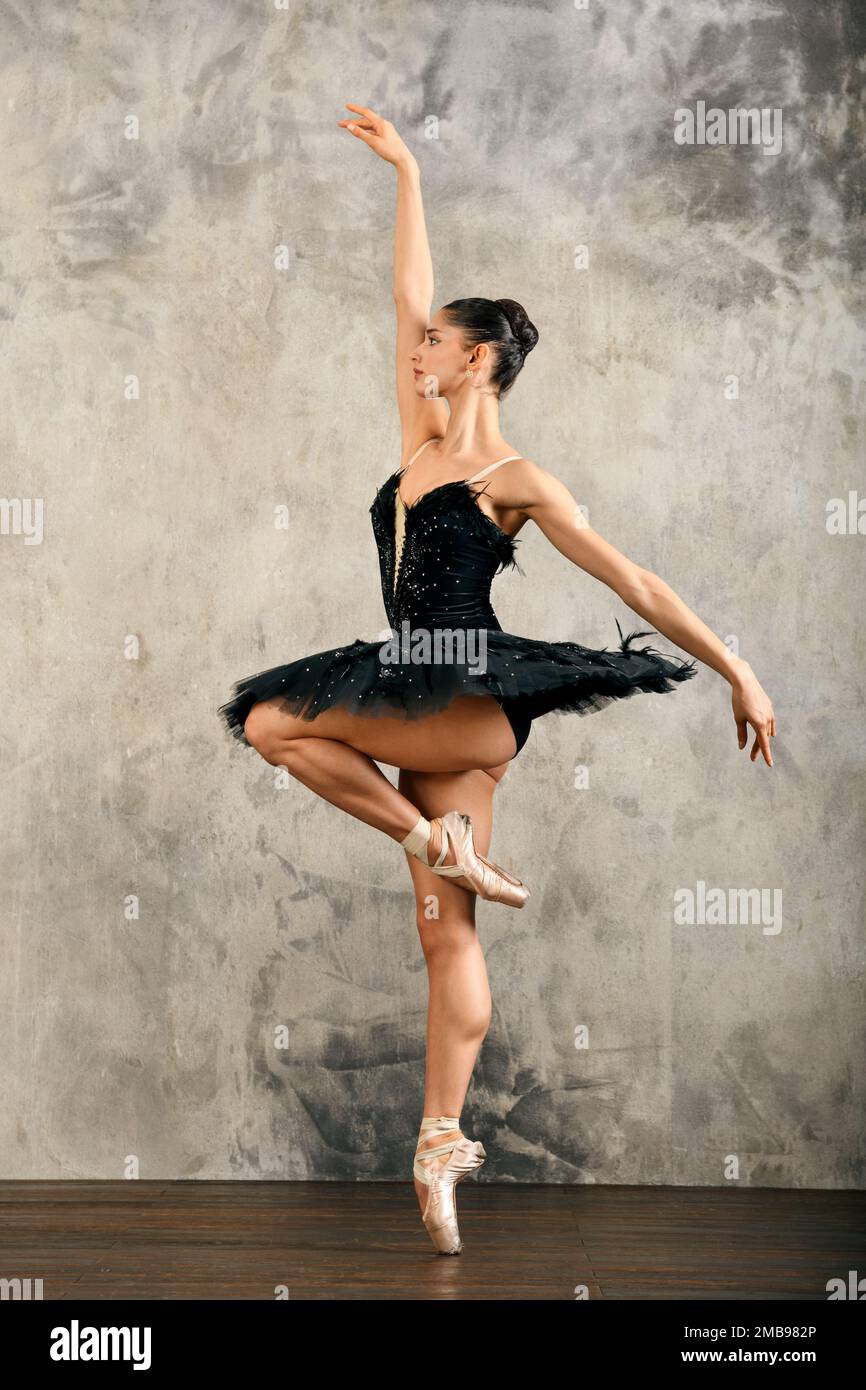 Vista laterale di una graziosa ballerina femminile con capelli scuri in gonna tutu nera e scarpe appuntite in piedi sul puntale con braccio rialzato mentre ballano contro g Foto Stock