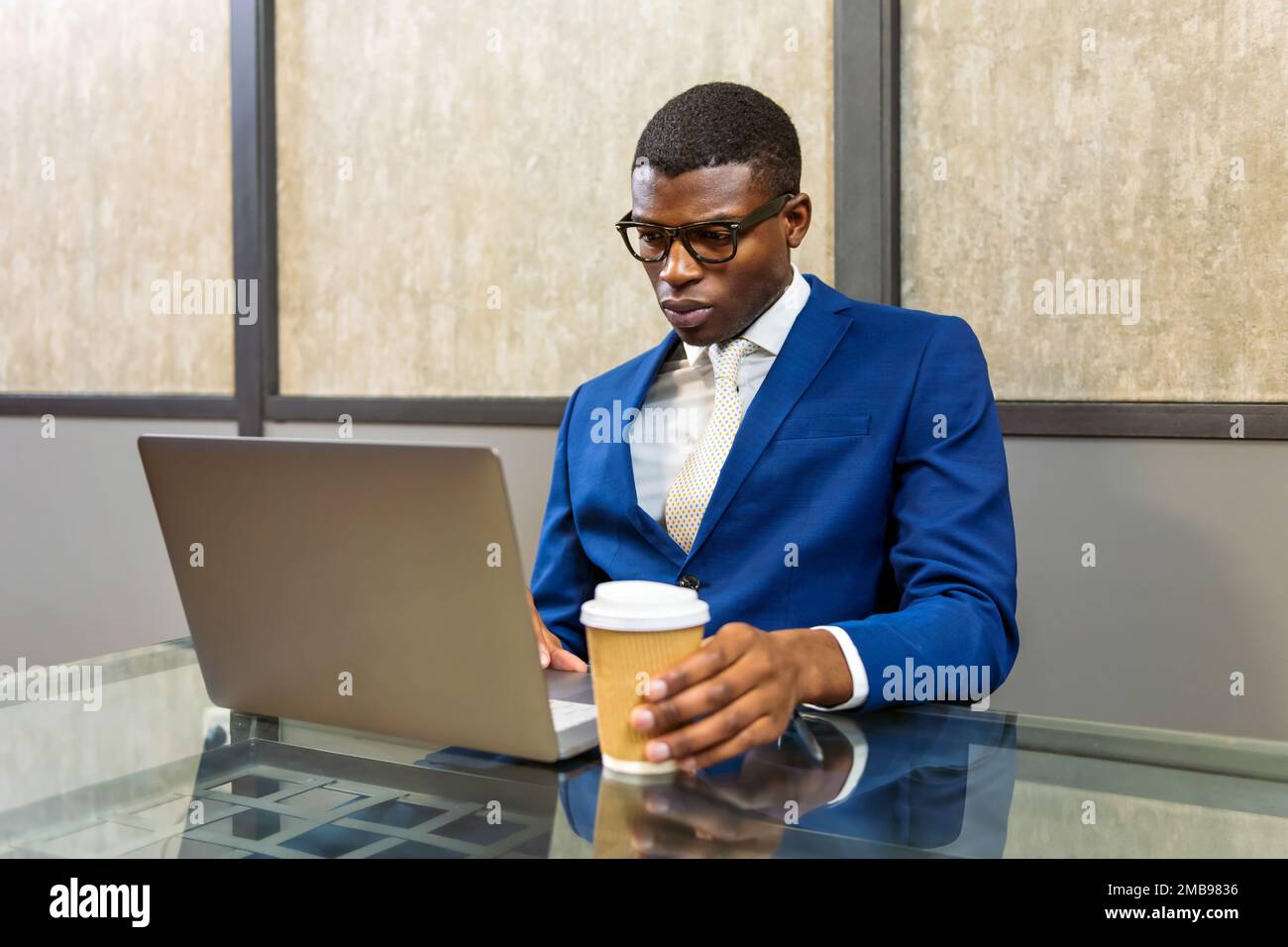 Serio impiegato afro-americano maschile in tuta blu formale e bicchieri con takeaway tazza di bevanda calda seduta a tavolo utilizzando netbook in ufficio Foto Stock